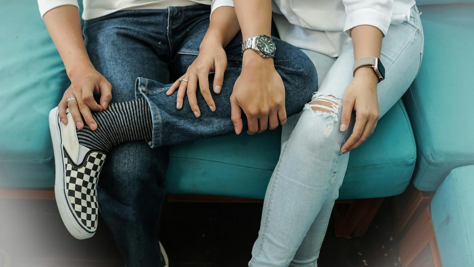 A young couple sitting comfortably on a teal sofa, showcasing casual fashion with denim and stylish watches.